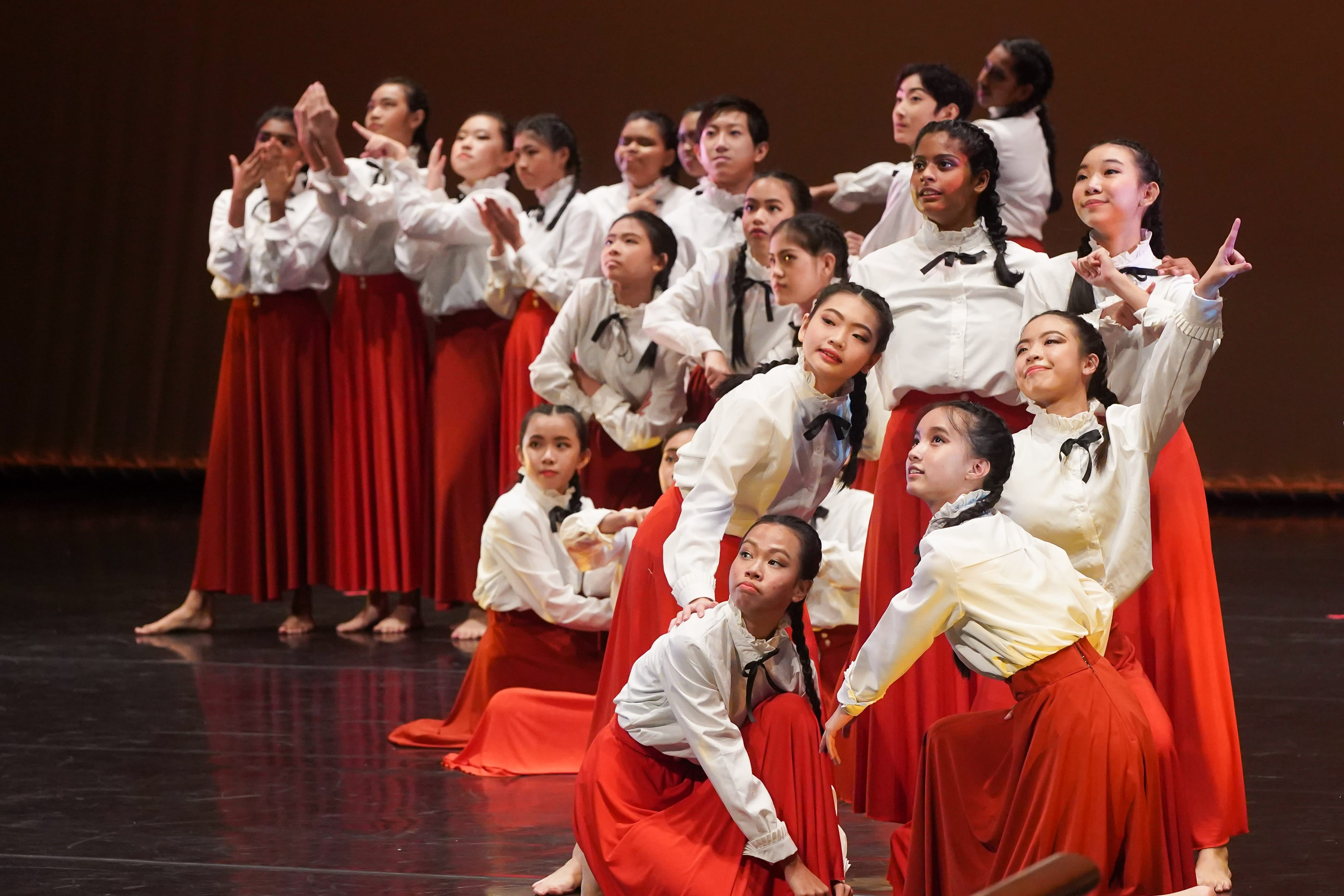 A large group of young female dancers in white blouses and red skirts performing on a dark stage.